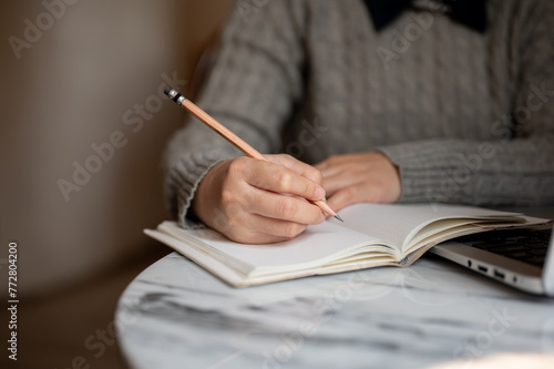 A young Asian female student is sitting indoors and writing on her notebook with a pencil.