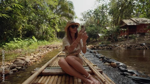 Female tourist floats on bamboo raft. Woman tourist enjoying the bamboo rafting on the river, films beautiful nature landscape on smartphone. Vacation, tropical tourism, travel, adventure lifestyle. photo