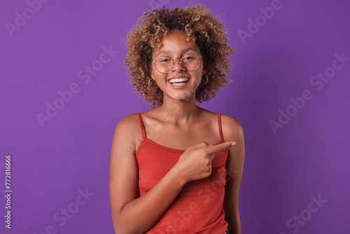 Young enthusiastic cute African American woman student with glasses points finger to side recommending pay attention to educational program for preparing for final exams stands in purple studio.