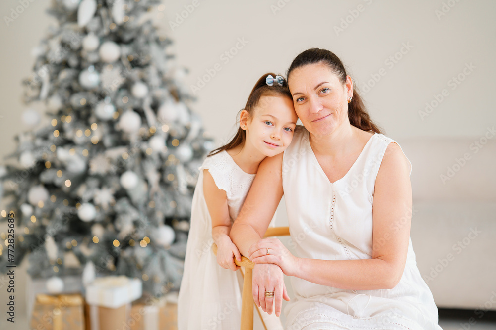 Gentle mother and daughter in white dresses at the Christmas tree. Happy family. Love and care. Gentle mother and daughter in white dresses at the Christmas tree. Happy family. Love and care.