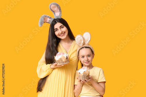 Happy smiling young woman with her daughter in bunny ears holding Easter cakes on orange background