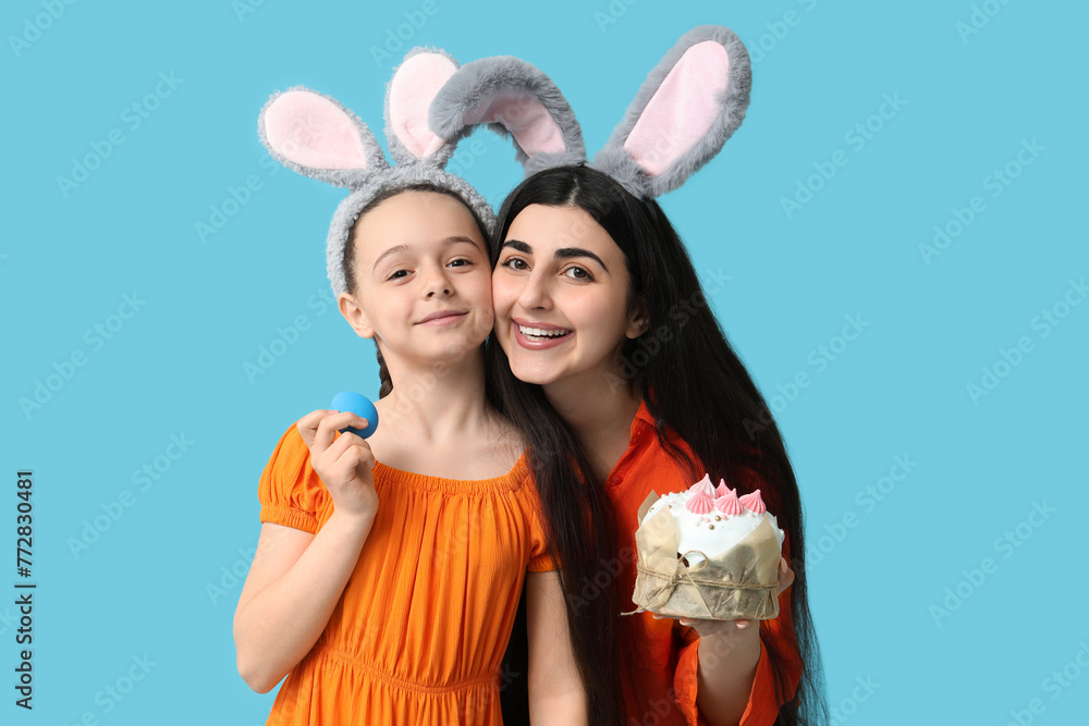 Happy smiling young woman with her daughter in bunny ears holding Easter cake and egg on blue background Happy smiling young woman with her daughter in bunny ears holding Easter cake and egg on blue background
