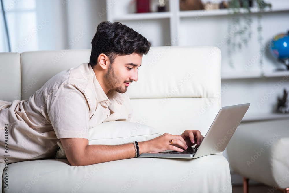 Relaxing Indian young man working on laptop while lying on sofa at home Relaxing Indian young man working on laptop while lying on sofa at home