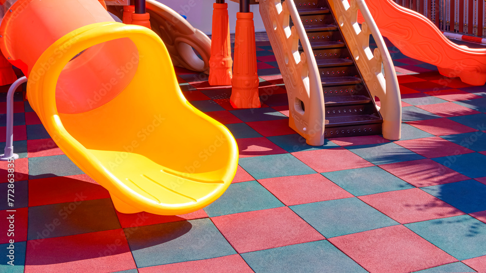 Slides with playground climbing equipment on colorful checkered rubber floor in outdoors playground area at kindergarten - obrazy, fototapety, plakaty 