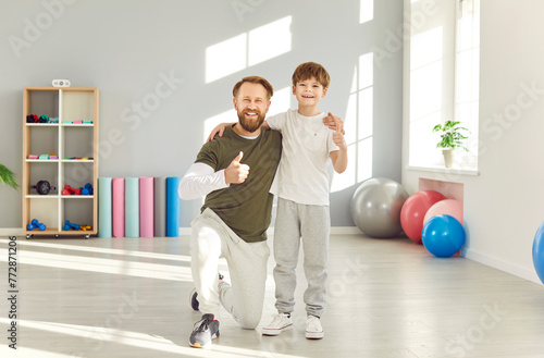 Portrait of a happy smiling father standing with his child boy in gym wearing sportswear looking cheerful at camera and showing thumb up sign after workout together. Family sport concept.