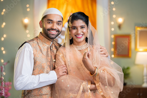 Happy smiling indian Muslim couple standing with embracing by looking at camera at home - concept of relationship, wishes and indian culture