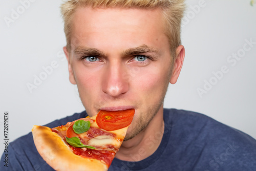 guy with pizza. a young guy with blond hair and a blue sweater sits on a white background and eats a piece of pizza  close-up  food concept