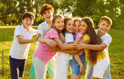 Portrait of happy smiling children friends standing on the green grass together in the park outdoors and looking cheerful at the camera hugging and having fun. Kids summer camp and holidays concept.