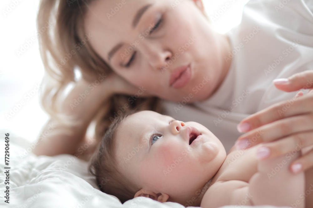 A 5-month-old baby and his beautiful blonde mom relax, play and laugh in bed in the bedroom. People a dressed in light home clothes, the family chooses natural textiles A 5-month-old baby and his beautiful blonde mom relax, play and laugh in bed in the bedroom. People a dressed in light home clothes, the family chooses natural textiles