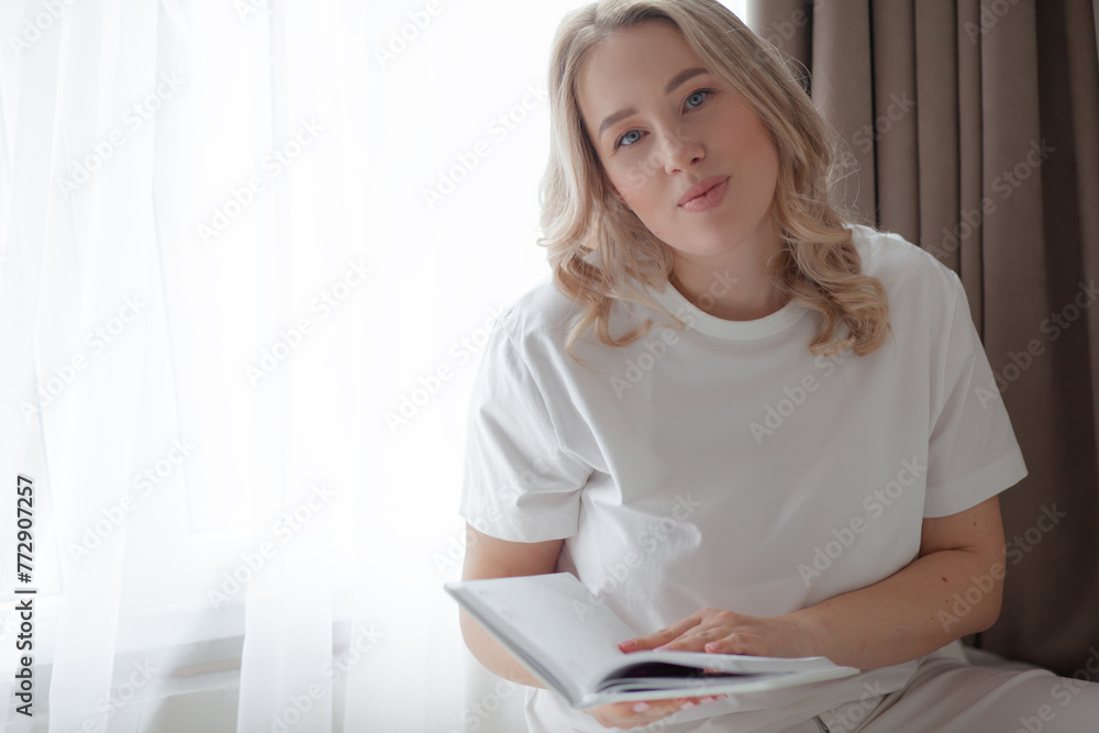 portrait of a beautiful European-looking blonde girl, dressed in light clothes, rest in n apartment against the background of windows. The woman holding notepad portrait of a beautiful European-looking blonde girl, dressed in light clothes, rest in n apartment against the background of windows. The woman holding notepad