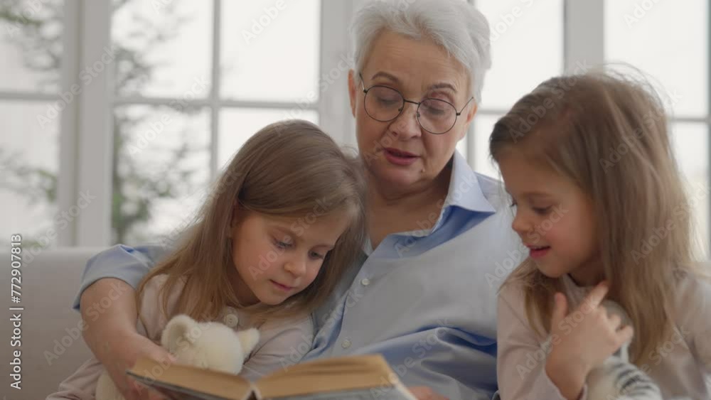 Grandmother with granddaughters resting. Serious relaxed older granny embracing reading book with children two twins granddaughters leisure rest on couch at home. Family love, generation concept. Grandmother with granddaughters resting. Serious relaxed older granny embracing reading book with children two twins granddaughters leisure rest on couch at home. Family love, generation concept.