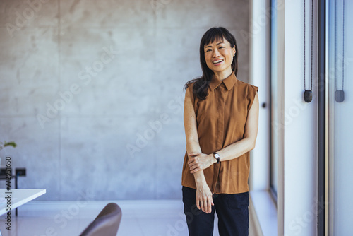 Portrait Of Female Designer Standing In Modern Office. Confident young Asian businesswoman standing smiling at the camera in a boardroom with colleagues in the background