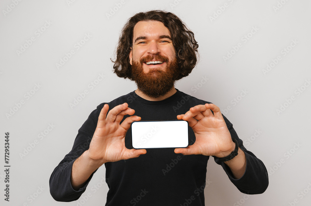 Studio shot of a smiling bearded man holding a phone with blank screen over white background. Studio shot of a smiling bearded man holding a phone with blank screen over white background.