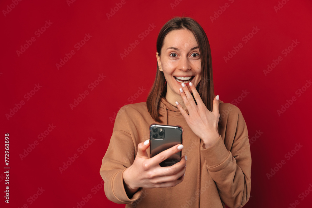 Excited shocked young woman is holding a phone over dark red background in a studio.