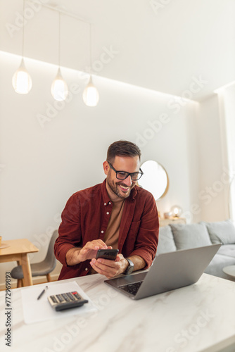 A smiling adult male accountant using his phone and a laptop while working from home