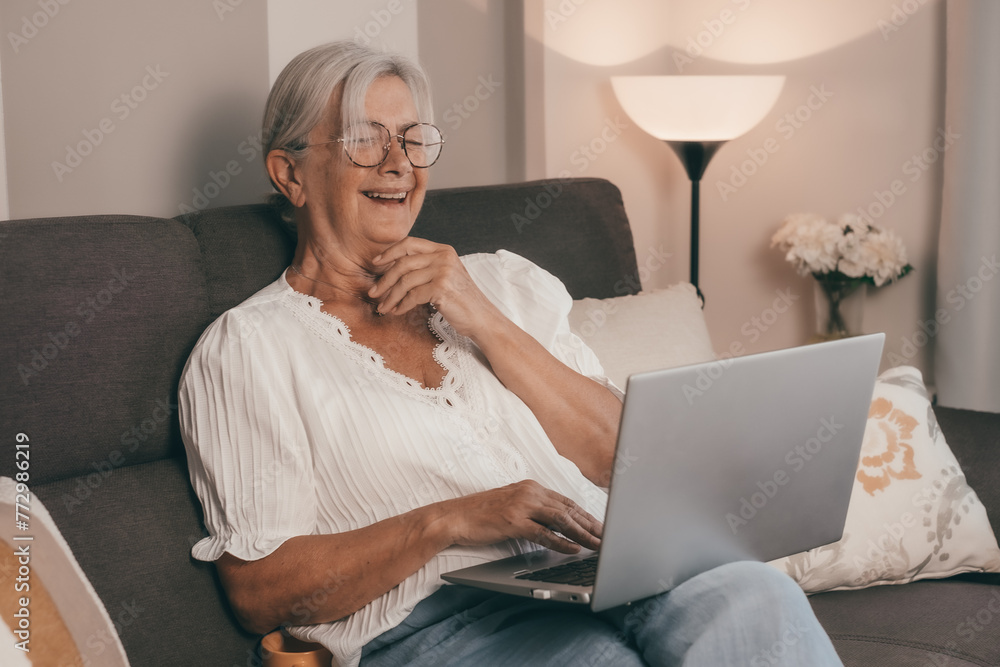 Happy amused senior white haired woman sitting on home sofa watching a funny movie on laptop enjoying tech and social Happy amused senior white haired woman sitting on home sofa watching a funny movie on laptop enjoying tech and social