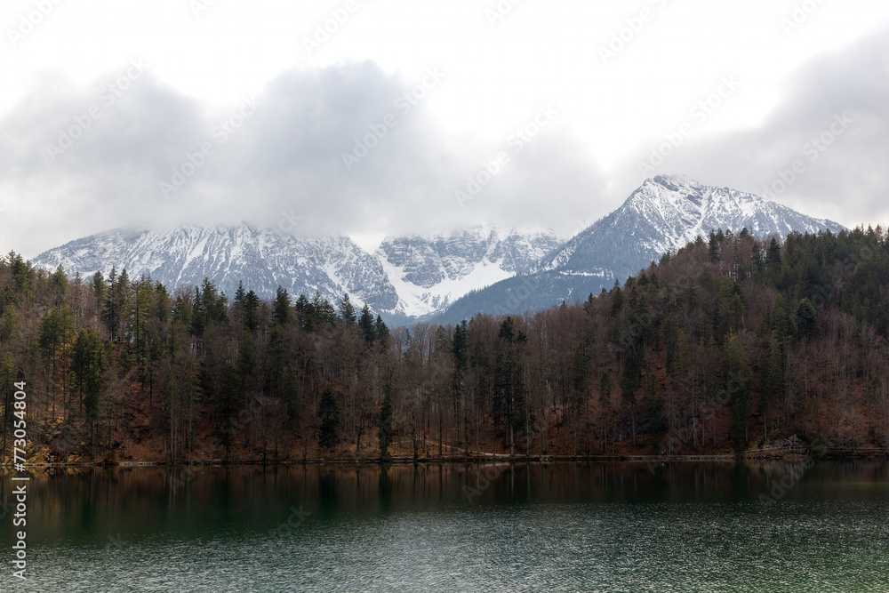 Blick in eine mit Wolken verdeckte, schneebedeckte  Bergkette am Ufer des Alatsee im Ostallgäu bei Füssen