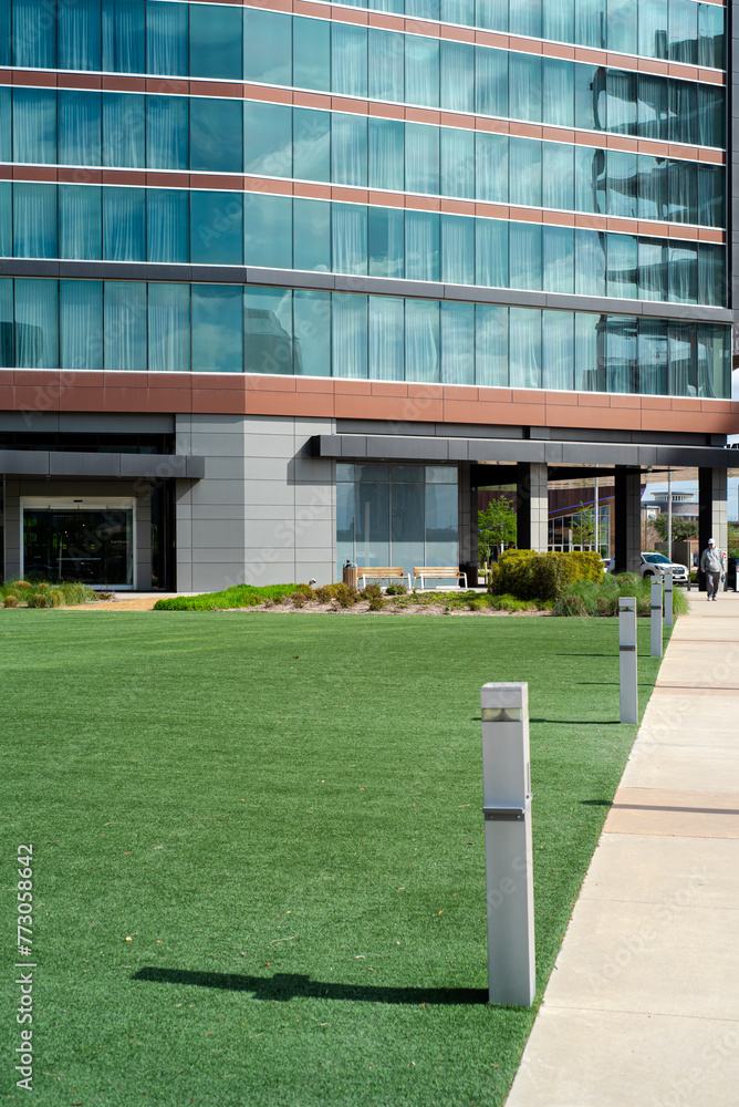 Concrete pathway with solar lights to entrance of multistory urban modern hotel glass wall blackout curtains in downtown Irving, Texas, large artificial grass courtyard, exterior building