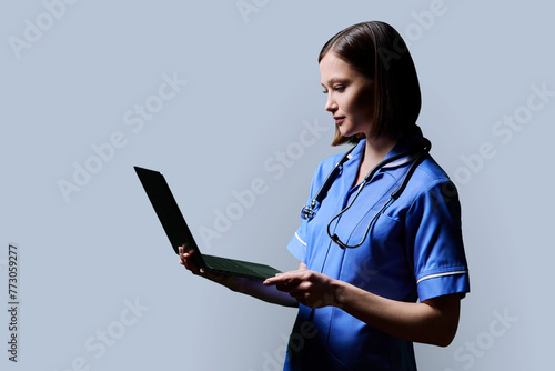 Female nurse using laptop, profile view on gray studio background