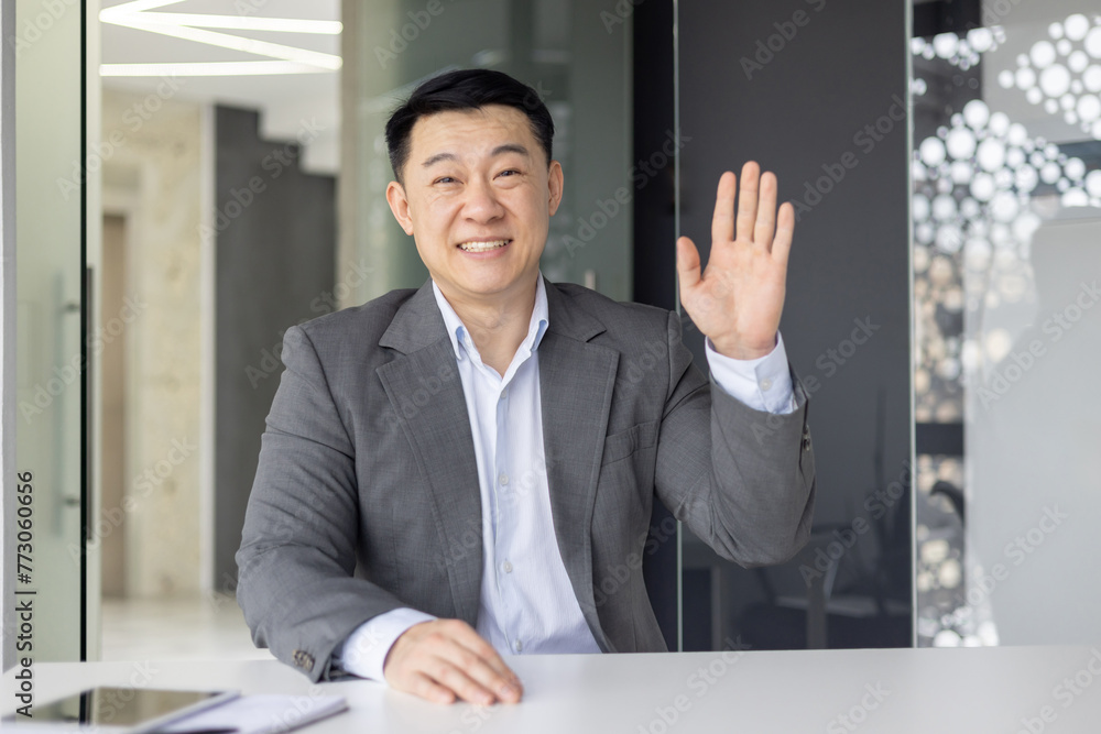 Professional Asian businessman waving hello while looking at the camera, engaged in a video conference call inside a modern office. Professional Asian businessman waving hello while looking at the camera, engaged in a video conference call inside a modern office.
