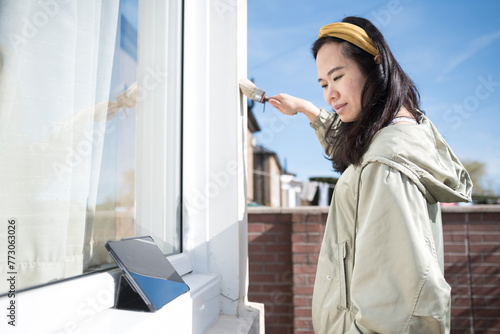 Asian woman wearing a yellow hairband paints the wall outside a window with a brush by watching an online tutorial on her digital tablet under a blue sky in Edinburgh, Scotland, United Kingdom