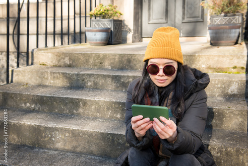 Young Asian woman wearing a yellow hat and scarf watching content on her green mobile phone in a sunny day while sitting on the steps at the front door of a house in Edinburgh, Scotland, UK
