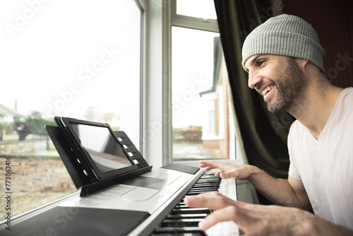 Young Spanish man wearing a grey hat plays piano and smiles while following an online piano lesson on his tablet sitting next to a bay window in Edinburgh, Scotland, UK