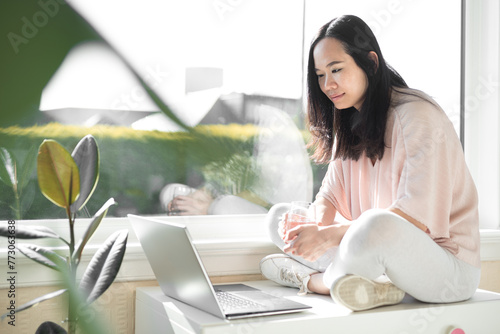 Young Chinese woman wearing a pink top and white trainers sits cross-legged on a white chest and smiles while watching content on her grey laptop computer next to a big bay window  and a plant
