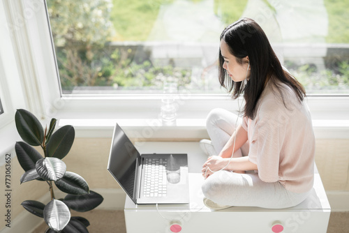 Asian woman wearing a pink top and white trainers sits cross legged on a white chest while watching content on her grey laptop computer and white earphones next to a big bay window and a plant