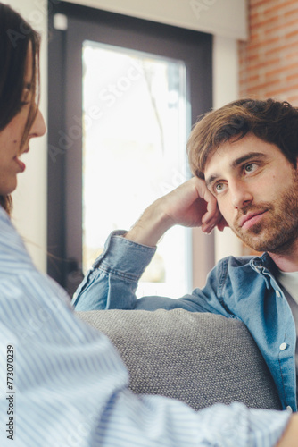 A young 25-year-old couple sits on their cozy sofa in a living room, engaged in a quiet conversation and sharing carefree moments together. Leisure and relaxation concept. Discussion man and woman