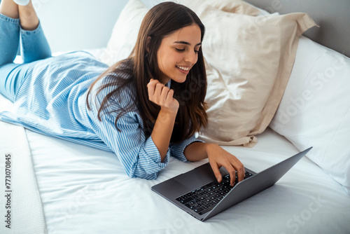A smiling woman lying down the bed in front of her laptop with her legs raised slightly. Working from home using computer and internet connection. Female people enjoy hotel room and remote work job