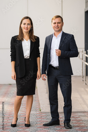Young business partners pose at camera for brilliant corporate photo. Businessman and businesswoman wear suits following dress-code of corporation