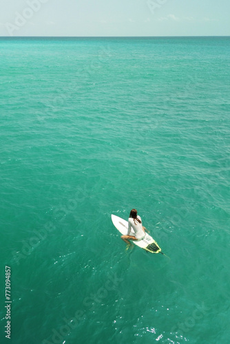 Vertical aerial view of a young sexy female surfer sitting on a surfboard on a line-up in a white swimsuit and waiting for a wave. Turquoise water in the ocean and big waves for surfing.
