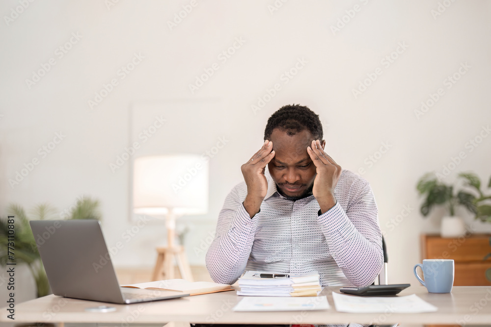 A young African businessman feels a headache. and stress while working on a laptop on a white wooden table in the office room A young African businessman feels a headache. and stress while working on a laptop on a white wooden table in the office room