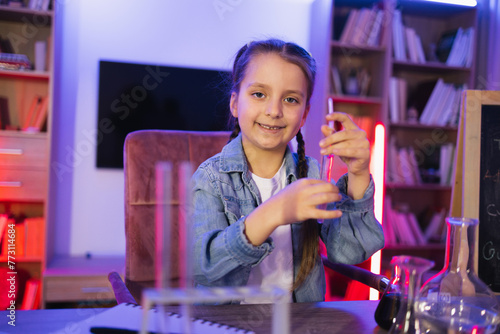 Science, hobbies, learning, education, discovery, childhood and domestic life. Focused Caucasian little girl doing chemistry experiment looking at test tube in evening at living room.