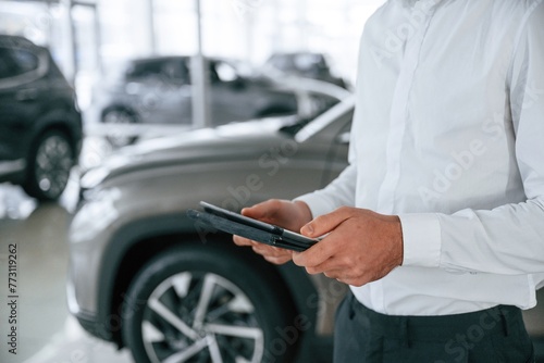 Black colored tablet in hands. Worker in formal clothes is in the auto salon