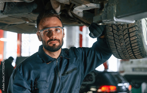 In protective eyewear, under vehicle. Car repairman is in the garage with automobile