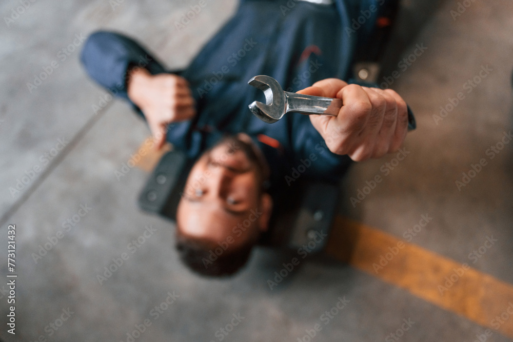 On the ground with tool. Car repairman is in the garage with automobile On the ground with tool. Car repairman is in the garage with automobile