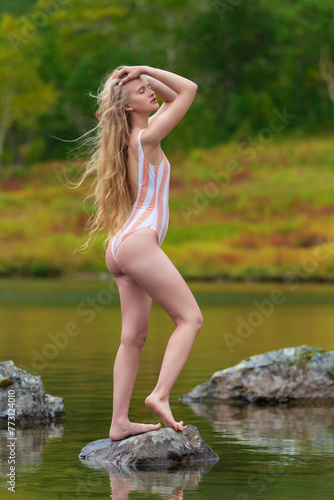 Woman with eyes closed in one piece swimsuit standing on stone rock island in middle of lake surrounded by autumn forest. Sensuality young woman looks so relaxed and happy, enjoying beauty of nature