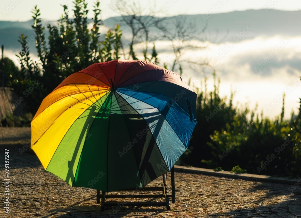 a rainbow umbrella is placed on an outdoor chair outside with mountains and trees behind it