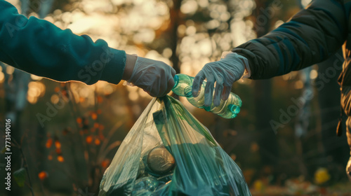 Gloved hands pass a plastic bottle over a full trash bag in a wooded area.