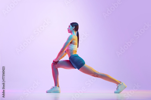 Young athlete woman doing lunges in motion in neon light against gradient studio background. Side view portrait. Concept of sport and recreation, movement, self care, action, energy. Ad
