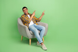 Full length portrait of positive guy sit cozy chair indicate fingers empty space offer isolated on green color background