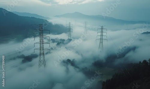 Aerial view of electricity transmission towers in a mountainous region, dense fog blanketing the ground with only the tops of the towers and mountains visible