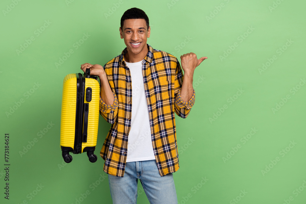 Photo of cheerful man hold suitcase indicate finger empty space offer isolated on green color background Photo of cheerful man hold suitcase indicate finger empty space offer isolated on green color background