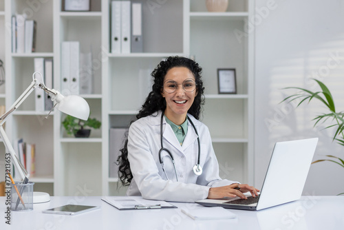 A cheerful Latino female doctor in a white coat smiles as she works on her laptop in a modern office setting. © Liubomir