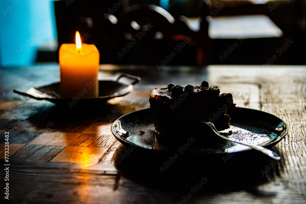 Burning wax candle and plate with a piece of berry chocolate cake and a fork on the wooden chessboard table. Cozy evening atmosphere in cafe, coffee shop. Dark blurred background. Selective focus. Burning wax candle and plate with a piece of berry chocolate cake and a fork on the wooden chessboard table. Cozy evening atmosphere in cafe, coffee shop. Dark blurred background. Selective focus.