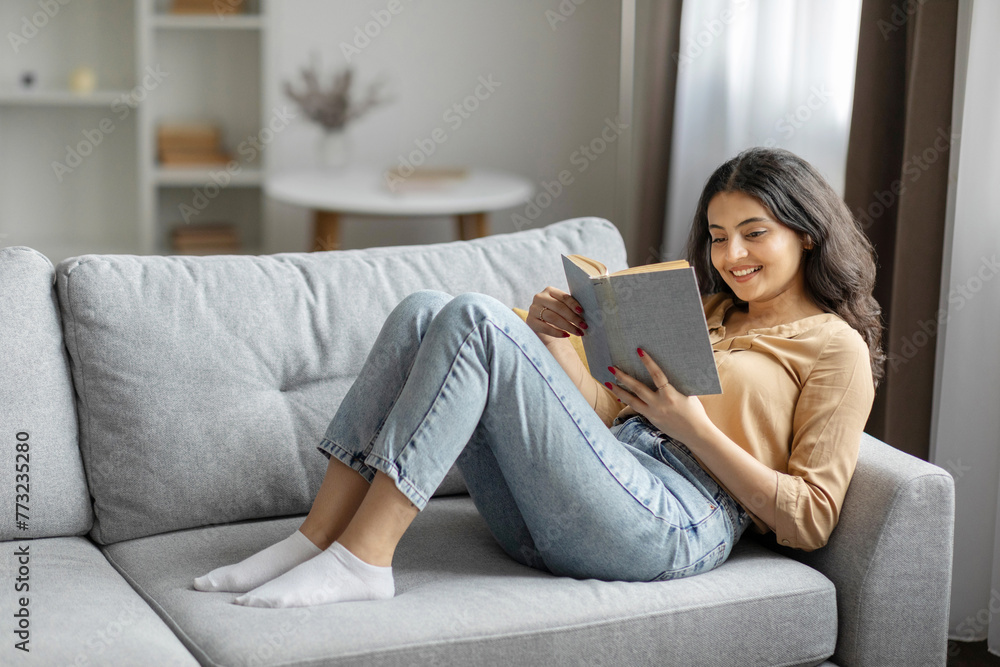 Young Indian woman lying on sofa with open book, enjoying peaceful morning, reading captivating story in living room, copy space Young Indian woman lying on sofa with open book, enjoying peaceful morning, reading captivating story in living room, copy space