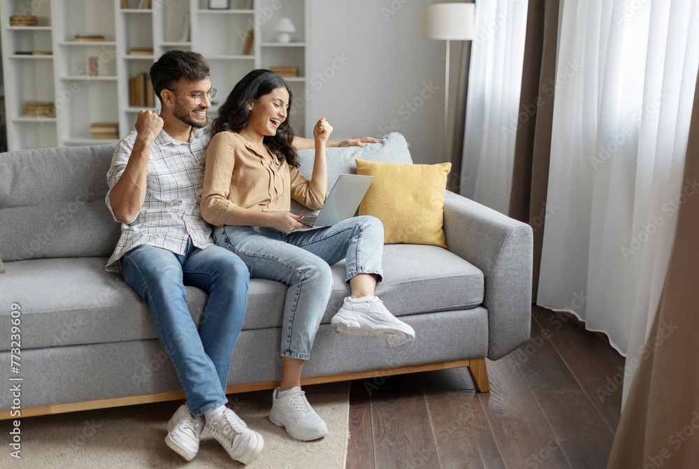 Emotional hindu couple with laptop making YES gesture, sitting on couch at home, celebrating success or enjoying sale in web store Emotional hindu couple with laptop making YES gesture, sitting on couch at home, celebrating success or enjoying sale in web store