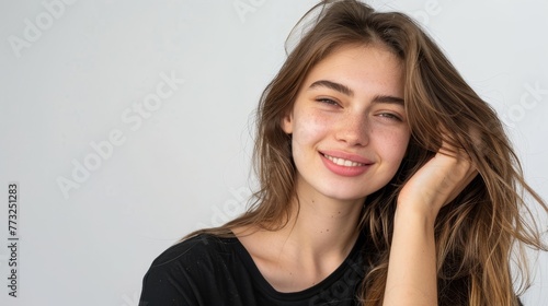 Stunning portrait of a beautiful young woman who is smiling, looking elegant and beautiful. She looks into the camera while sitting isolated on a white background wearing a black t-shirt and touching